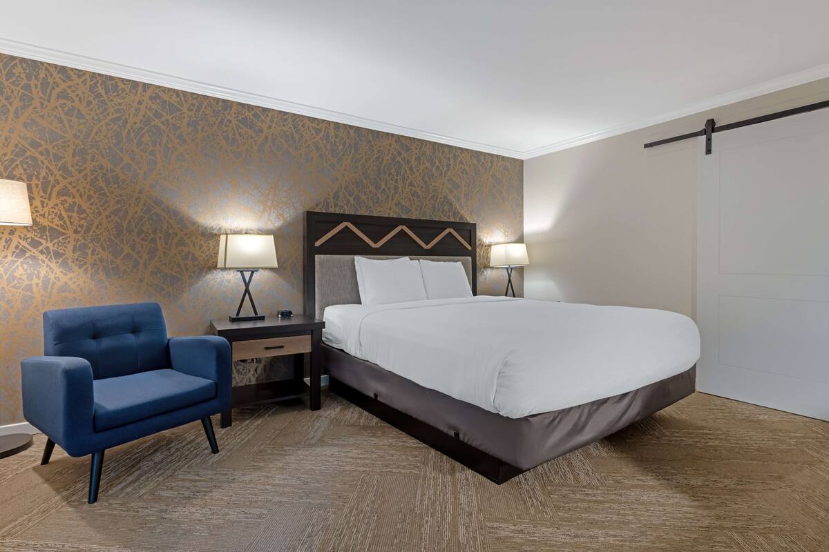A hotel room with a large bed, a blue armchair, two bedside tables with lamps, and a patterned wall.