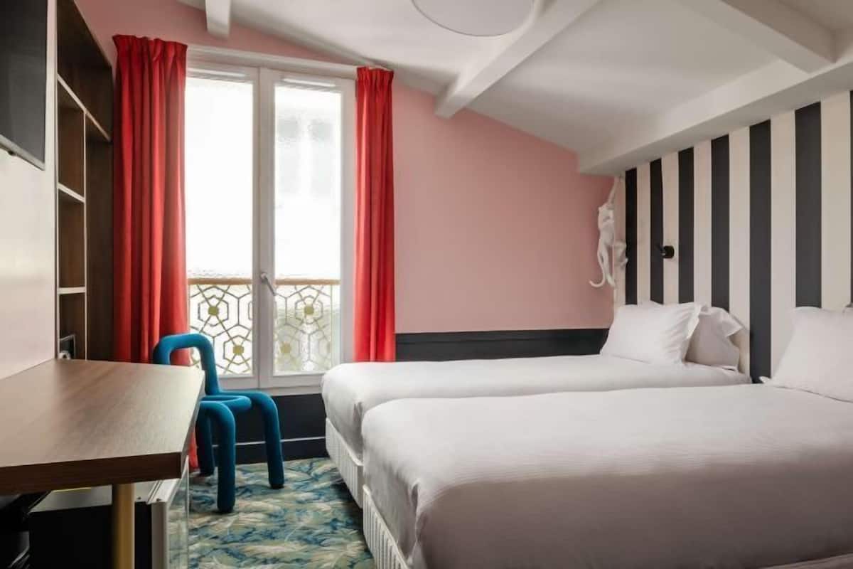 Quarto de hotel com duas camas, uma mesa, uma cadeira e uma janela com cortinas.