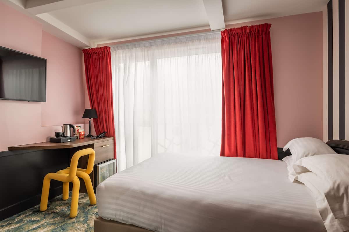 Quarto de hotel com uma cama, uma escrivaninha com cadeira, uma televisão e uma janela com cortinas vermelhas.