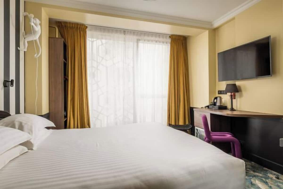 Quarto de hotel com cama, escrivaninha, cadeira, televisão e guarda-roupa.
