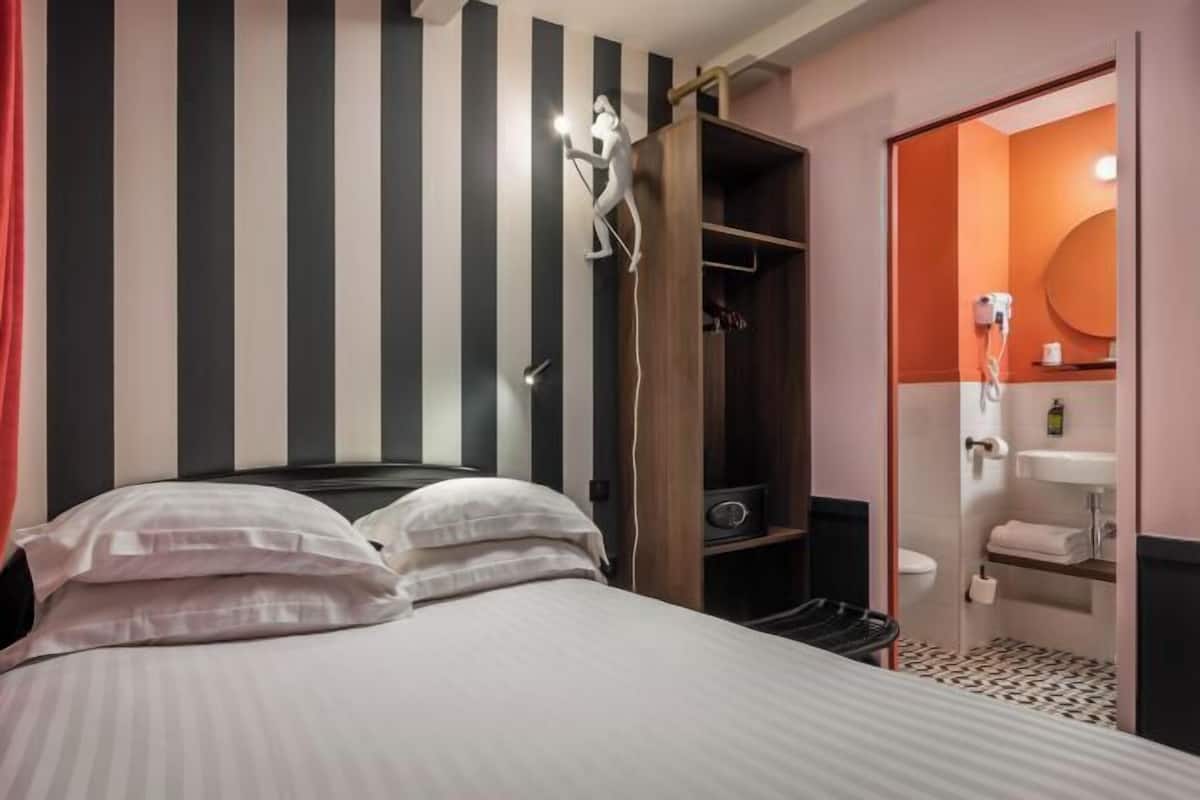 Quarto de hotel com parede listrada, cama com roupa de cama branca, guarda-roupa, banheiro com pia e espelho, e cortina vermelha.