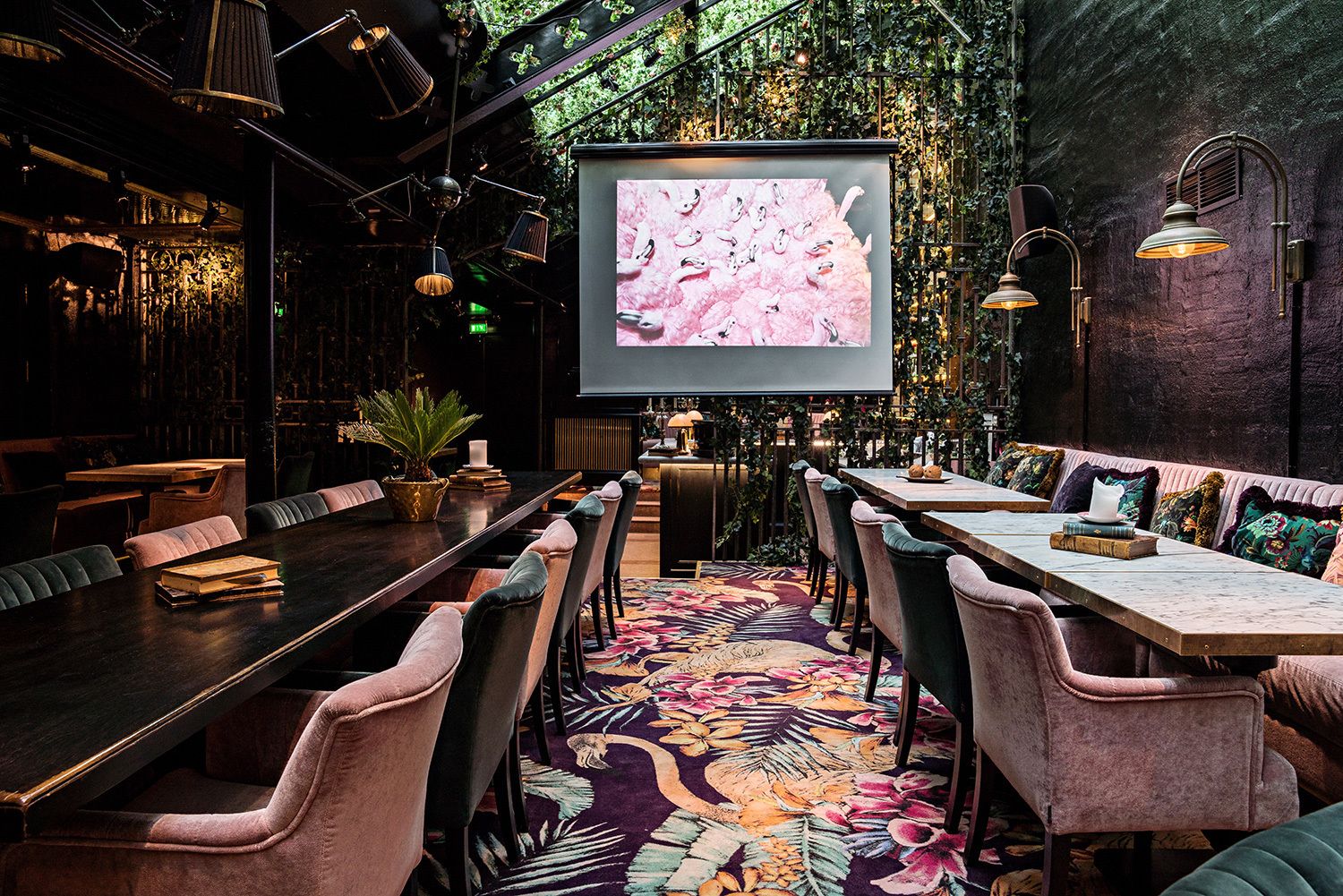 A cozy restaurant with a long communal table, cushioned chairs, and a large screen displaying a pink floral pattern.