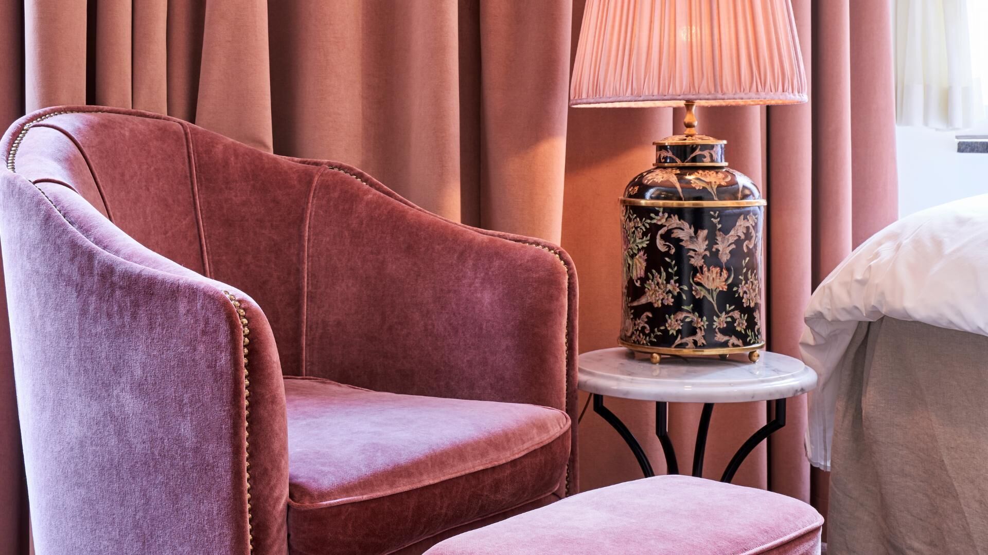 En blød, rød lænestol og skammel i et hotelværelse, med en dekorativ lampe på et lille bord ved siden af stolen.