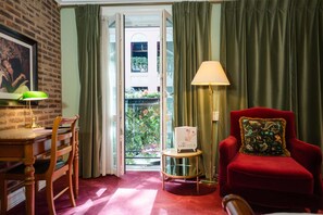 A room with a wooden desk, a red armchair with a decorative pillow, a green lamp, and a view of a balcony with plants.