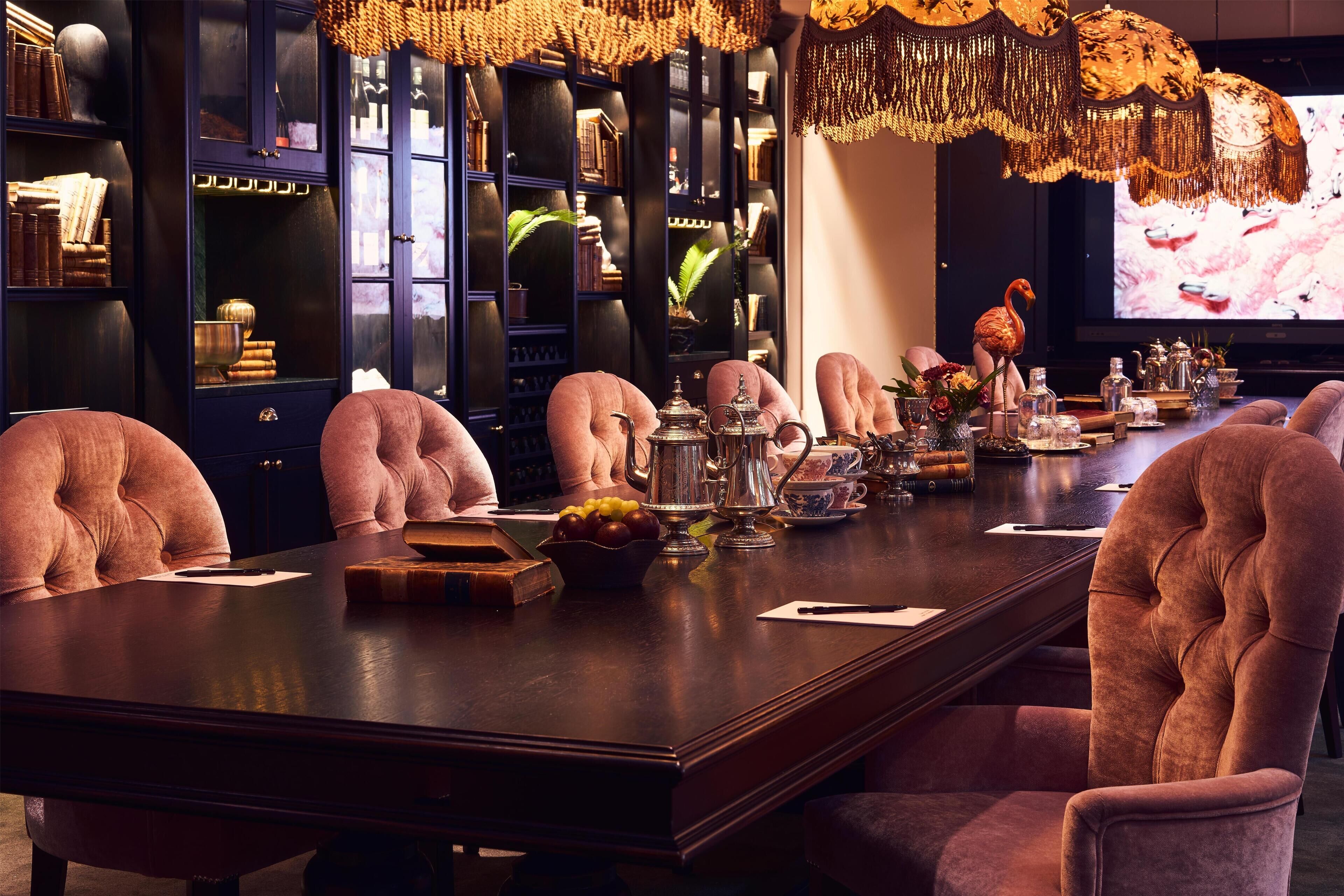 A long dining table with pink upholstered chairs, surrounded by dark wood cabinets with glass doors, and a television screen in the background.