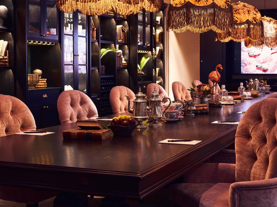 A long dining table with pink upholstered chairs, surrounded by dark wood cabinets with glass doors, and a television screen in the background.