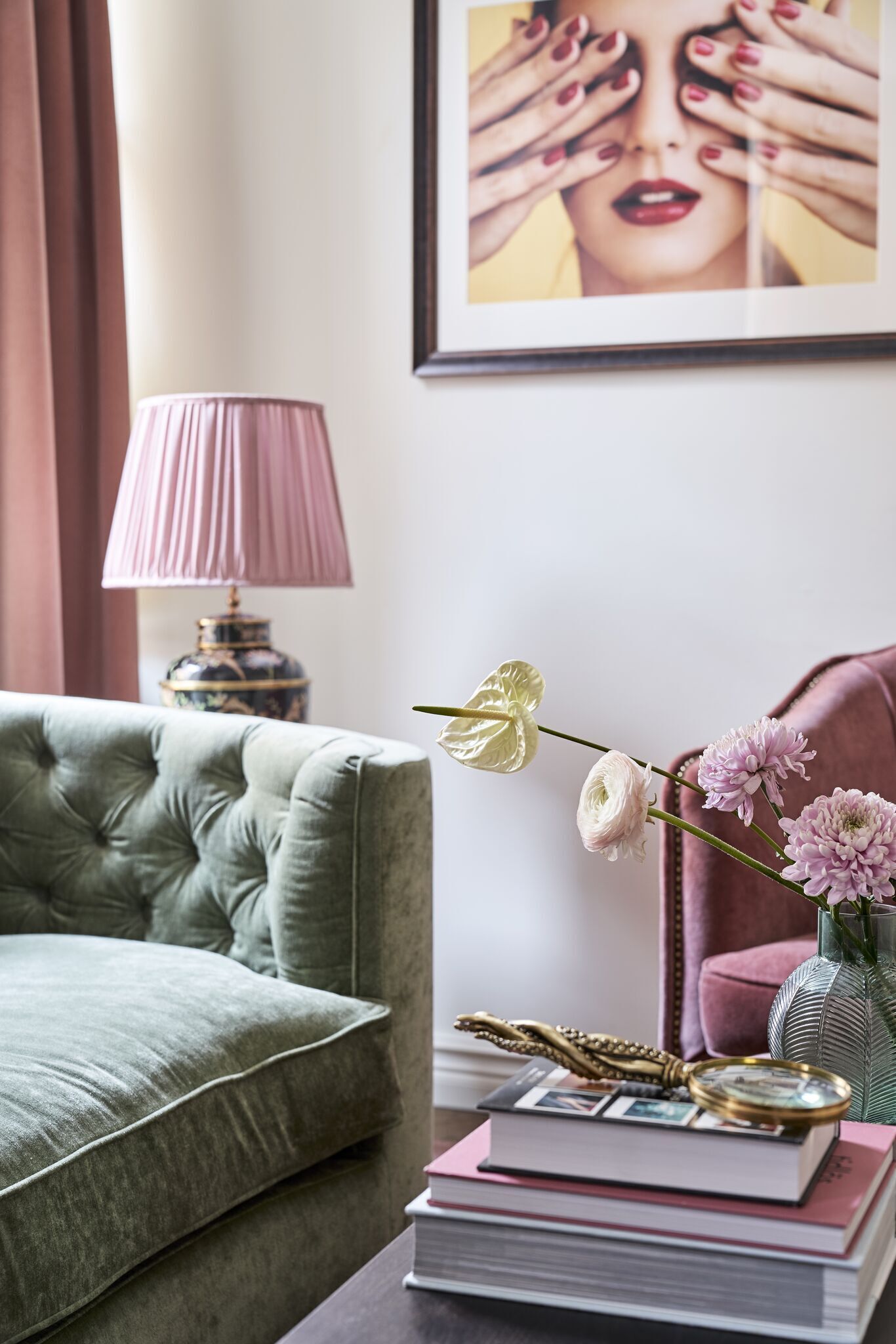 A green sofa, a pink lamp, a framed picture, a vase with flowers, and a stack of books.