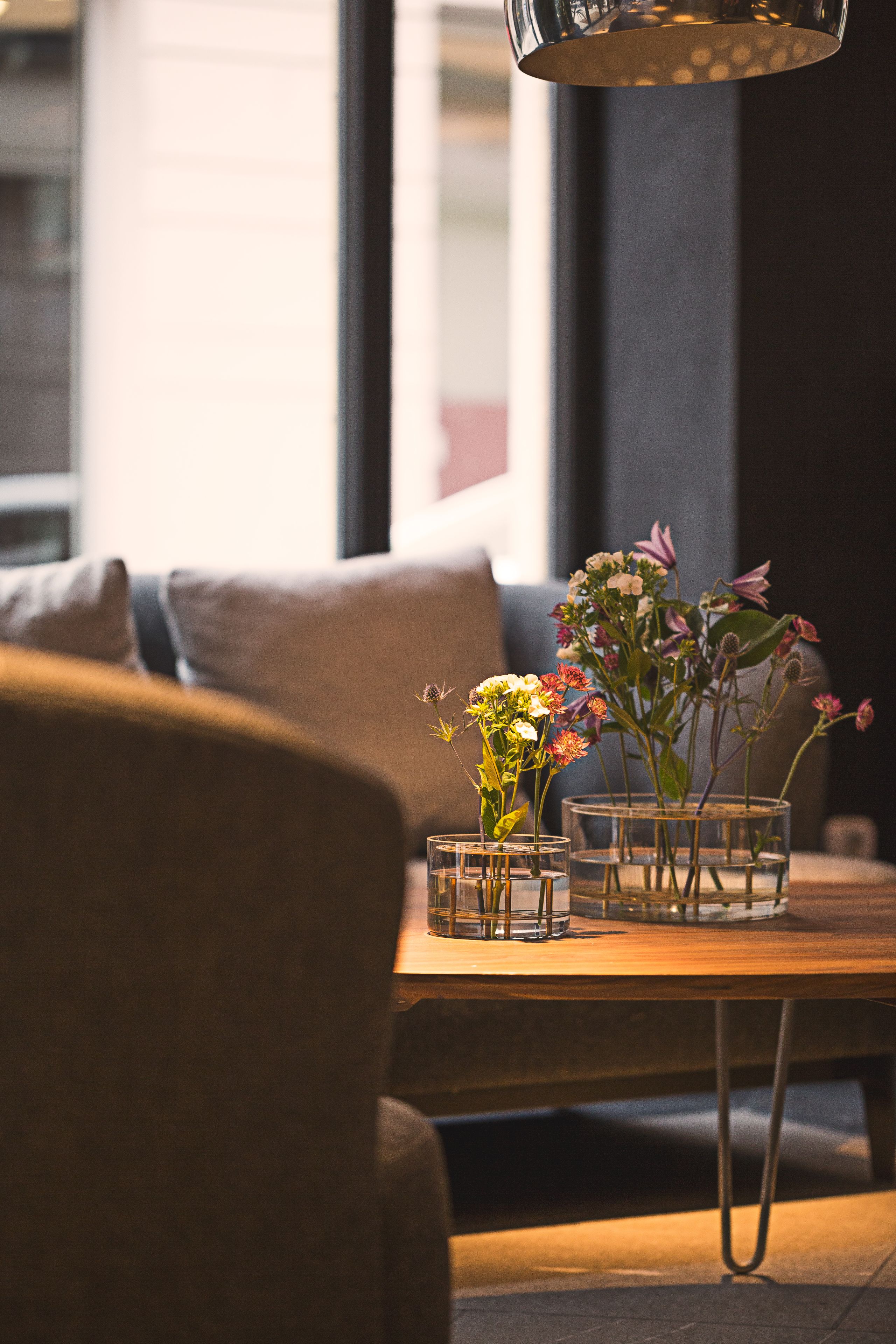 A modern living room with a sofa, a wooden coffee table with glass vases of flowers, and a hanging light fixture.
