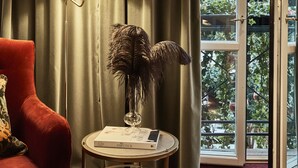 A room with a red armchair, a lamp, a small table with a book and a decorative item, and a view through a glass door to an outdoor area with plants. - MJ's (Malmö)