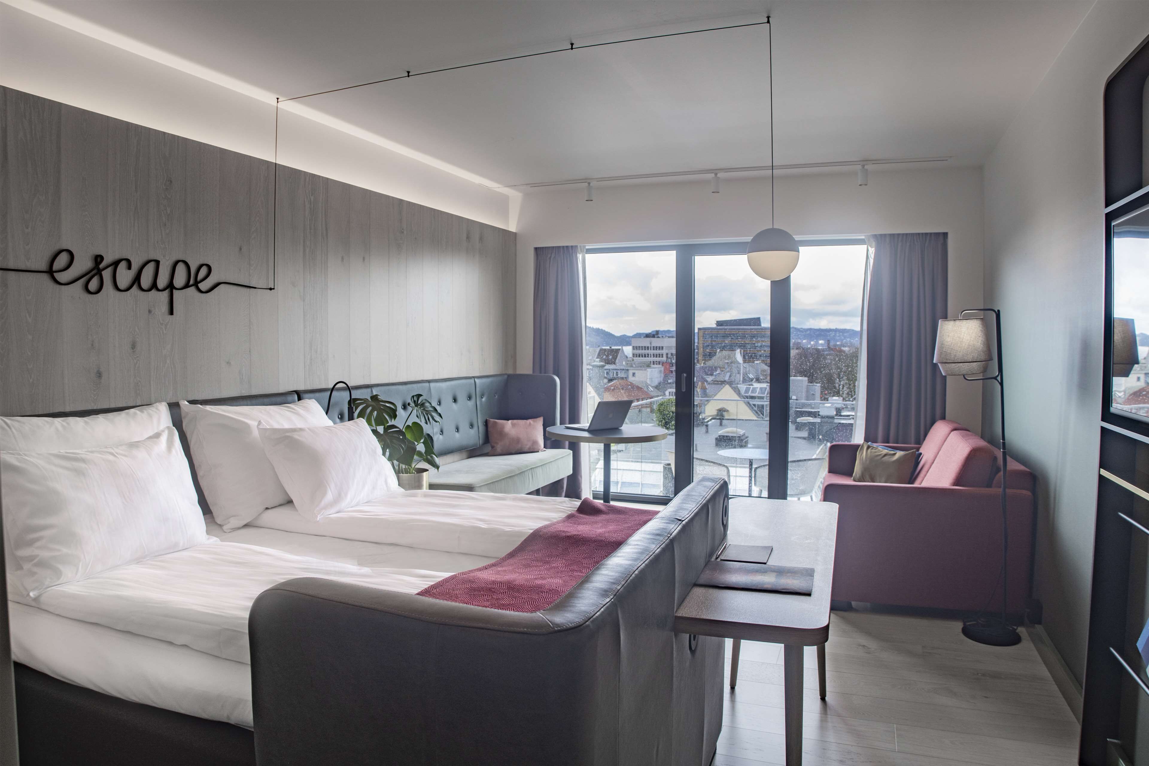 A modern hotel room with a large bed, a sofa, a small table, and a view of the city through large windows.