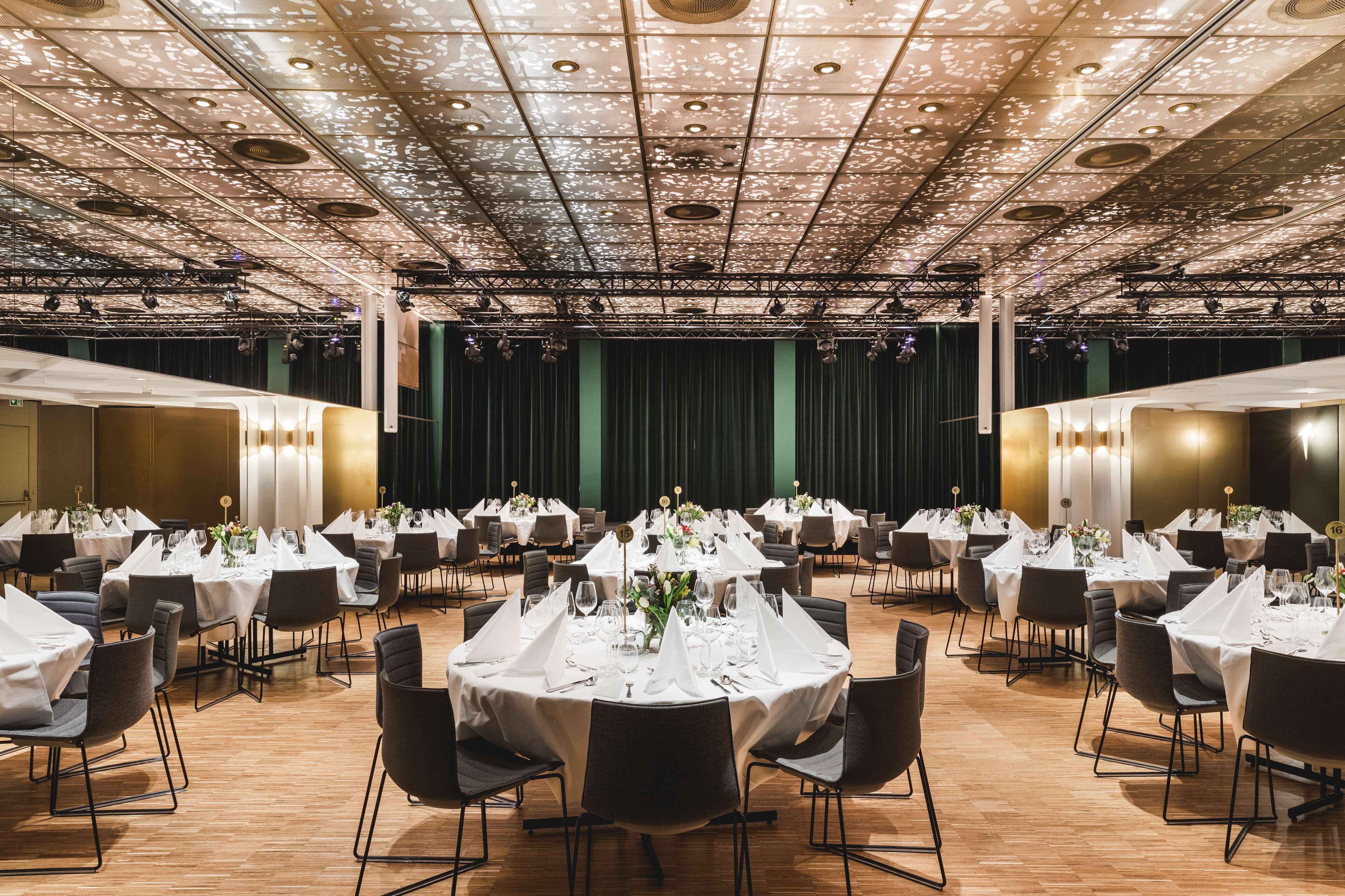 A banquet hall with round tables set for dining, a stage with curtains, and a patterned ceiling.