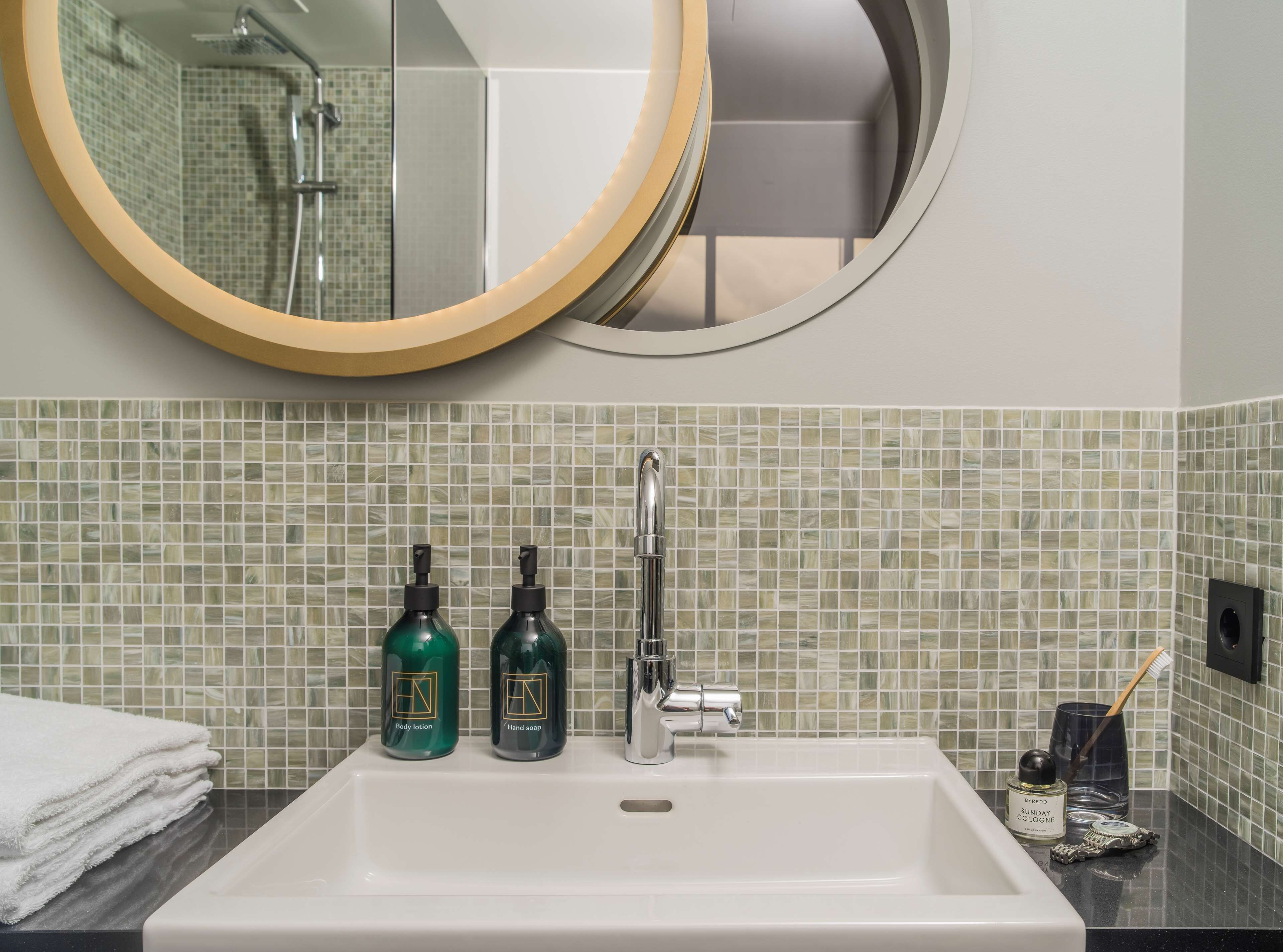 Un baño con lavamanos blanco, dos botellas de loción, un espejo y una ducha.