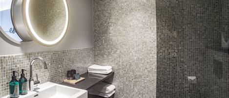 Un baño moderno con un lavamanos blanco, un espejo con luz circular y una zona de ducha con azulejos.