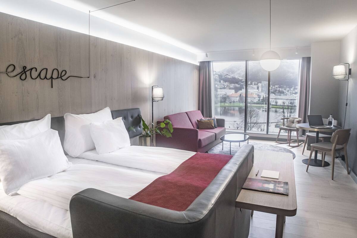 A modern hotel room with a large bed, a sofa, a desk, and a view of a cityscape through large windows.