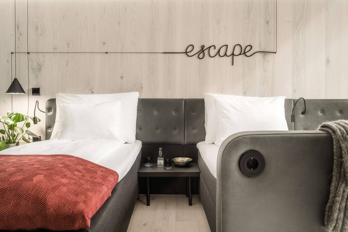 A modern bedroom with a grey upholstered bed, a bedside table with a lamp, and a wall-mounted word "escape".
