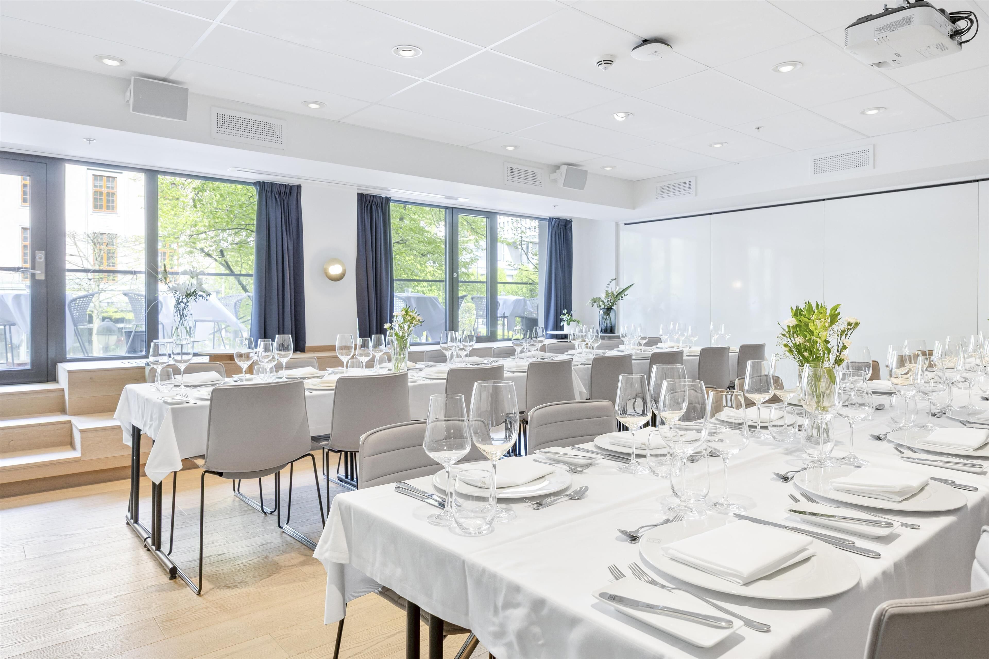 Un salón de conferencias con mesas dispuestas para un evento formal, ventanas amplias y una pantalla de proyector.