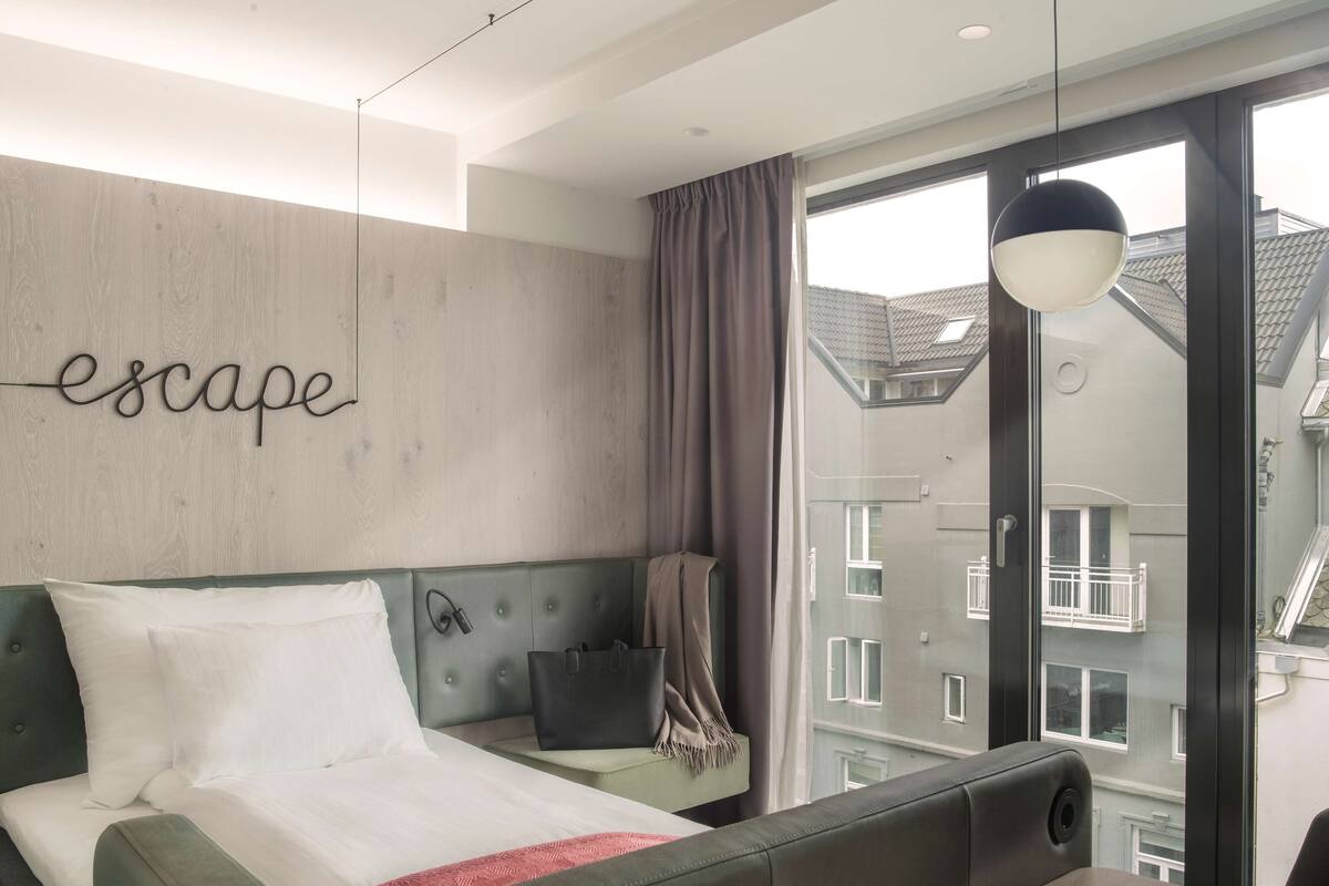A modern hotel room with a bed, a bedside table, a lamp, and a view of the cityscape through large windows.