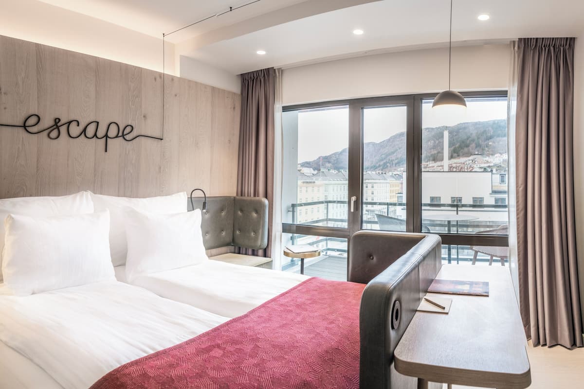 A modern hotel room with a large bed, a desk, and a view of a cityscape through large windows.
