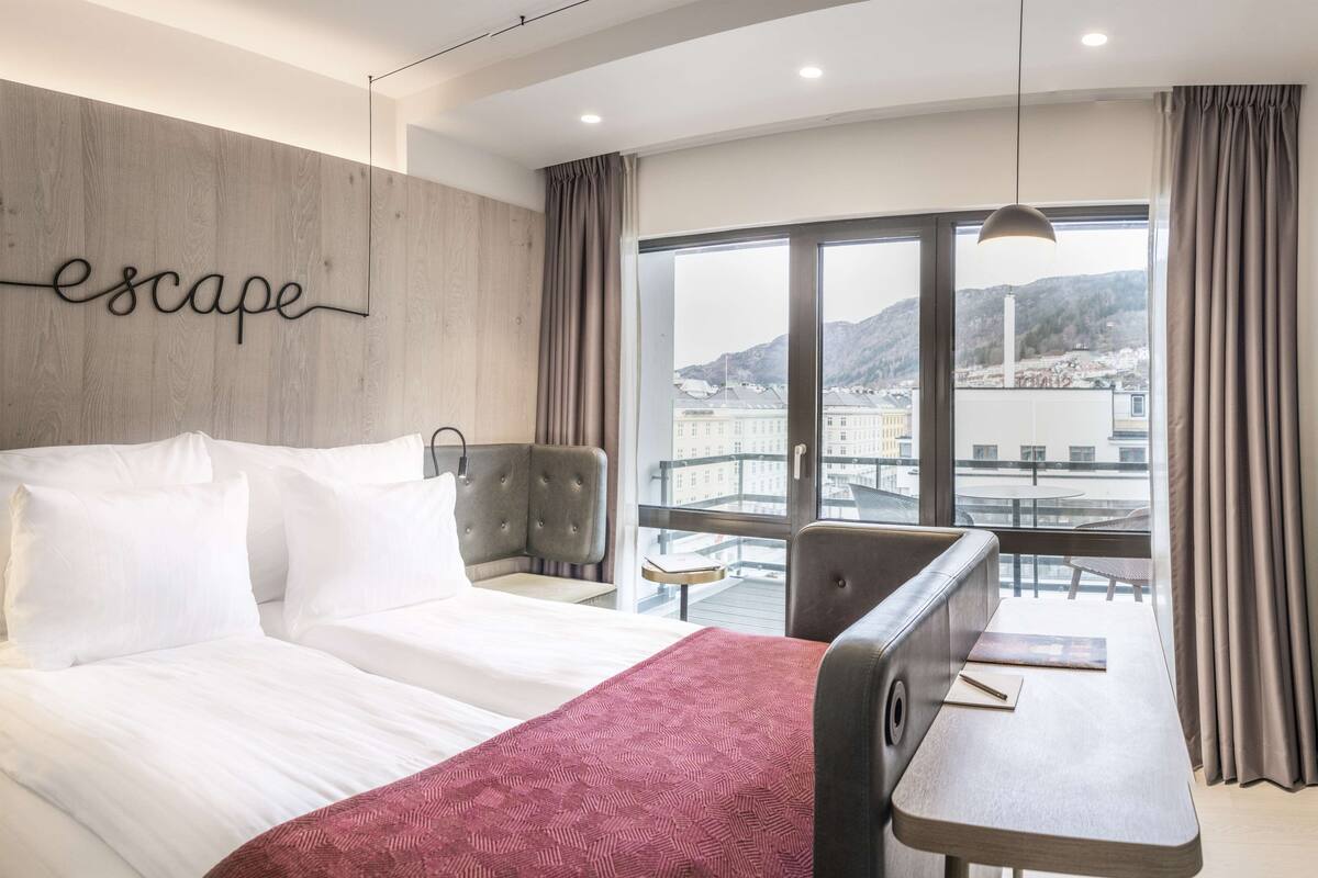 A hotel room with a bed, a desk, and a view of a cityscape through large windows.