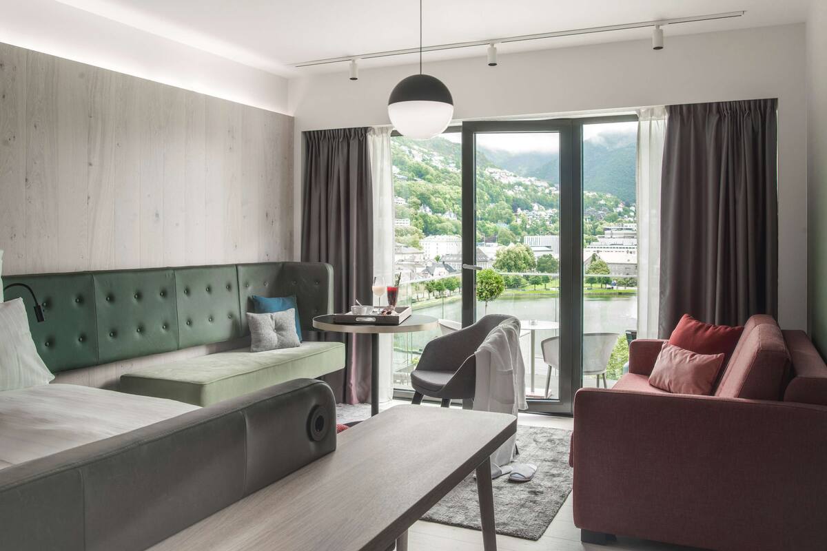 A hotel room with a bed, a green sofa, a red sofa, a small table, and a view of a city and mountains through large windows.