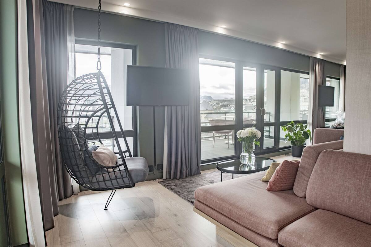 A modern living room with a hanging chair, a sofa, a flat-screen TV, and large windows offering a city view.