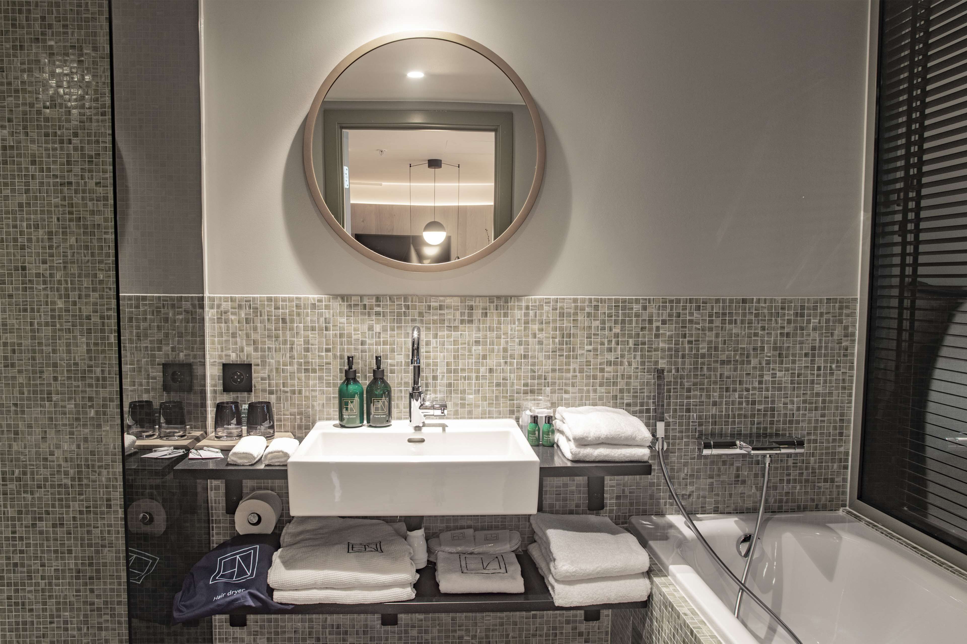 Un baño moderno con un espejo redondo, lavamanos blanco y salpicadero de azulejos. Hay toallas y artículos de aseo en la encimera.