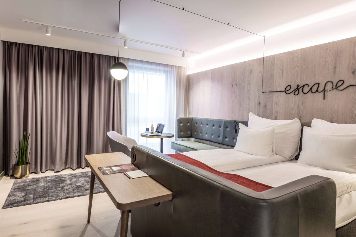 A modern hotel room with a sofa bed, a small table, a potted plant, and a wall with the word "escape" written on it.