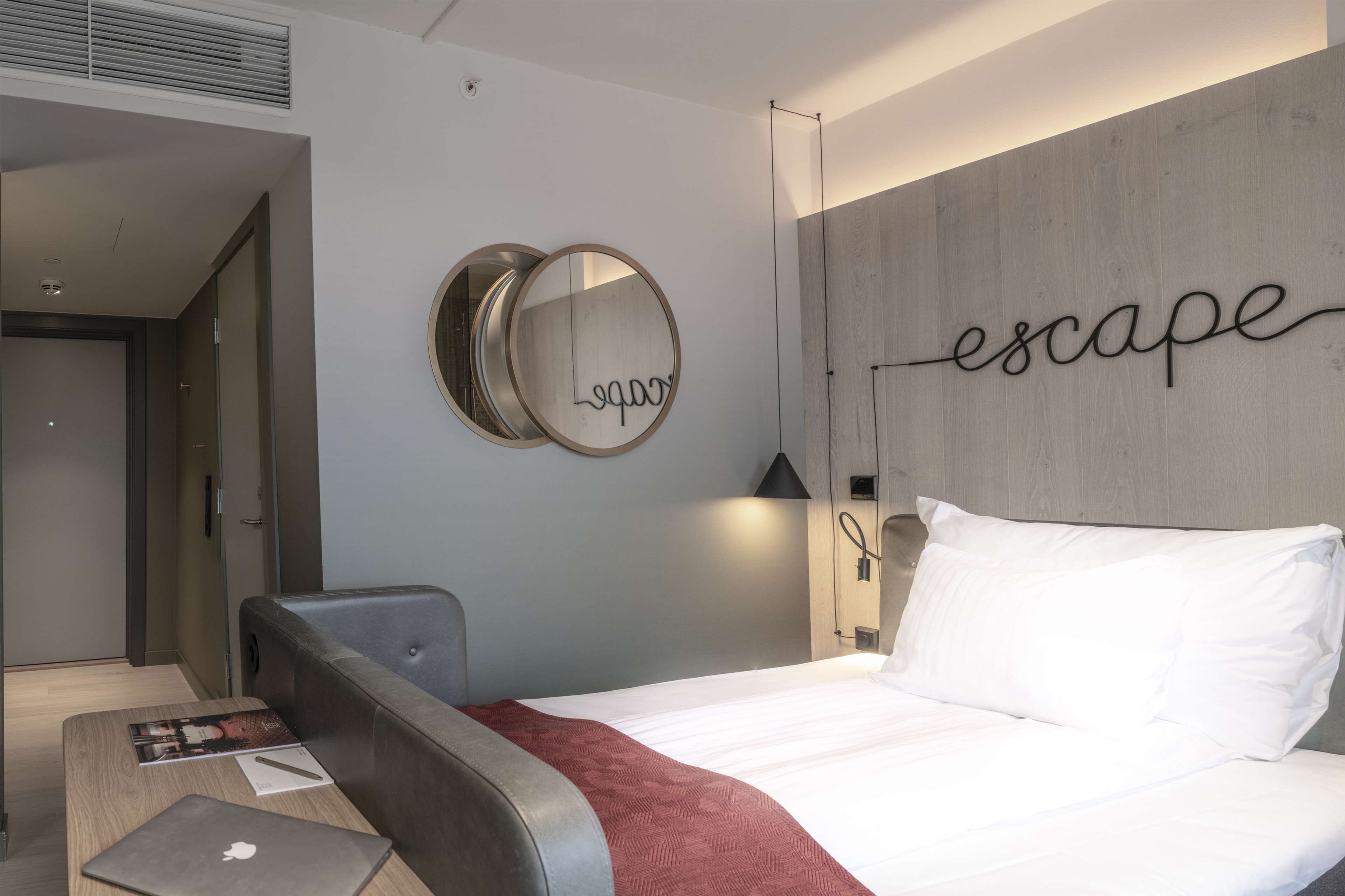 Una habitación de hotel moderna con una cama, un escritorio con una computadora portátil, un espejo y una pared con la palabra 