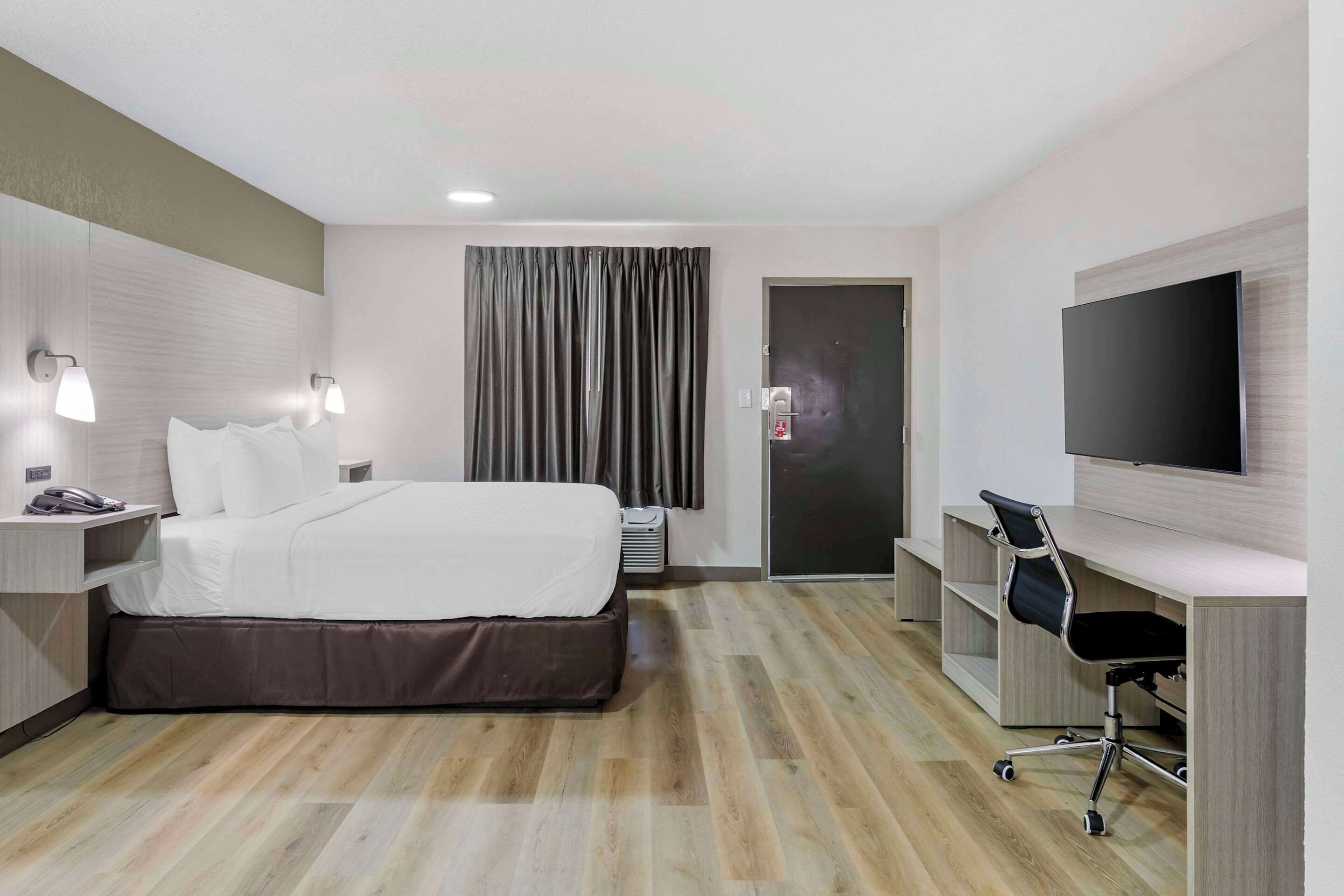 A hotel room with a bed, bedside table, lamp, desk, chair, TV, and a door.