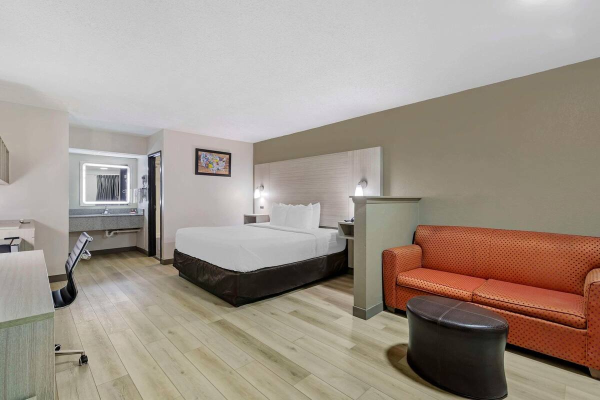 Una habitación de hotel con cama, escritorio, silla, sofá naranja y un cubo de basura.