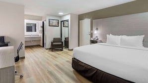 A hotel room with a large bed, a desk with a chair, a vanity with a mirror, and a door leading to another room.