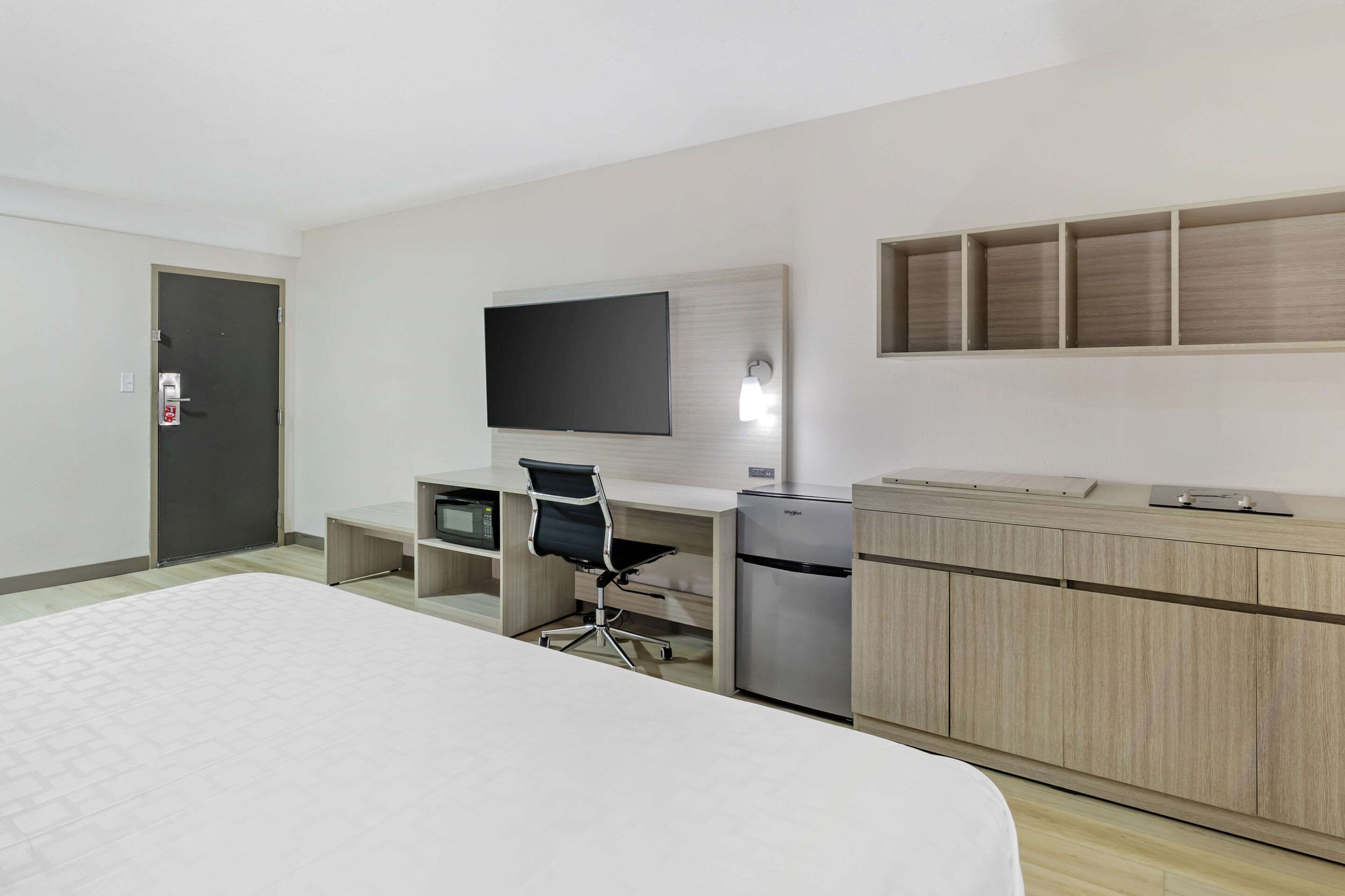 A hotel room with a bed, a desk with a computer, a refrigerator, and a TV mounted on the wall.