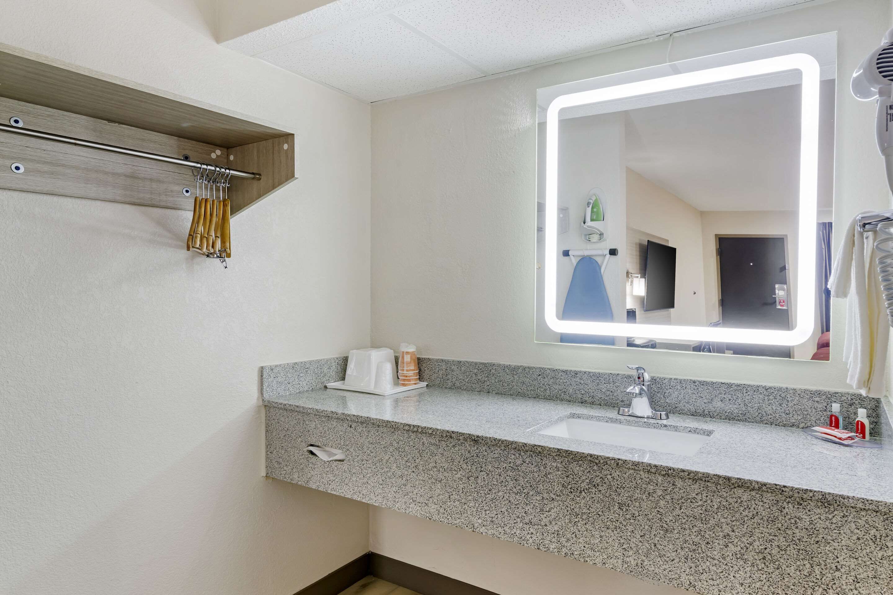 A modern bathroom with a granite countertop, a mirror with integrated lighting, a towel rack, and a small shelf with toiletries.