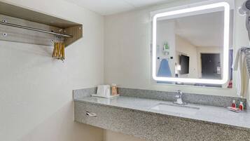 A modern bathroom with a granite countertop, a mirror with integrated lighting, a towel rack, and a small shelf with toiletries.