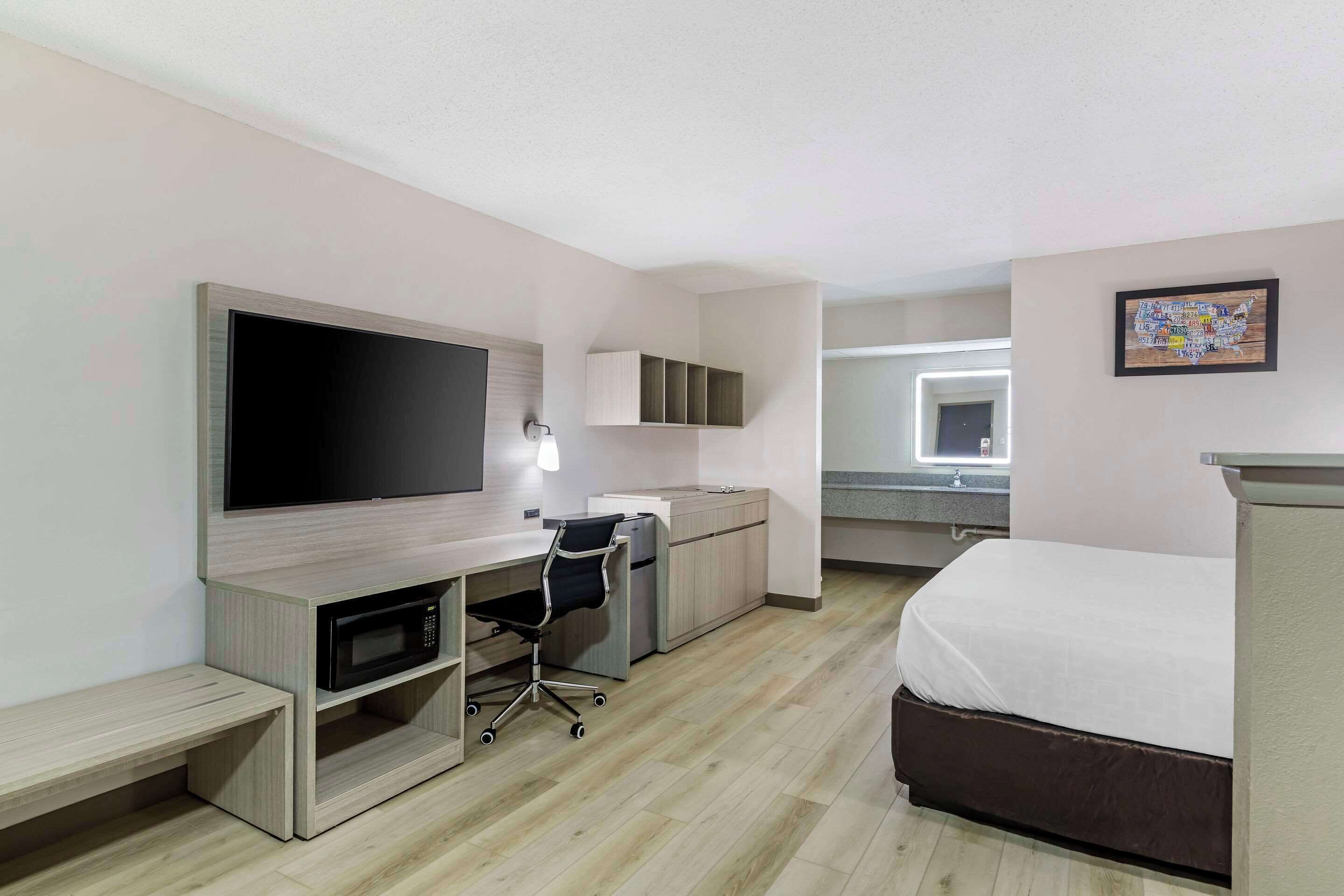 Una habitación de hotel moderna con una cama grande, un escritorio con computadora, un televisor y una pequeña cocina.