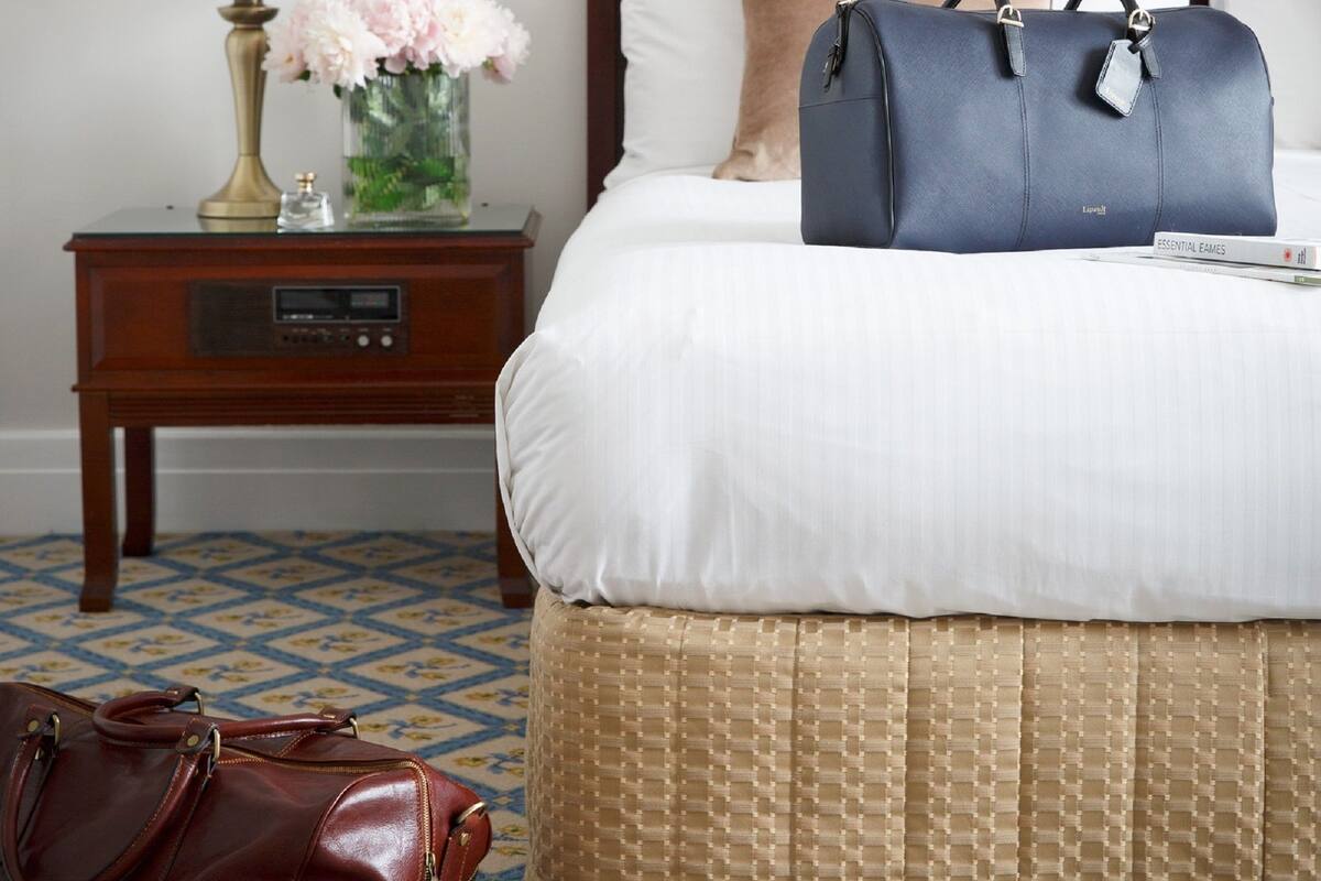 A neatly made bed with a blue bag on top, a brown bag on the floor, and a nightstand with a lamp and flowers.