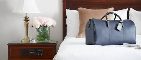 Una cama bien hecha con un bolso azul encima, un bolso marrón en el suelo y una mesita de noche con una lámpara y flores.