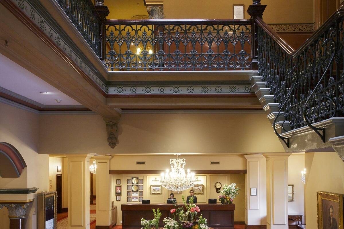 A grand lobby with a central reception desk, a chandelier, and a staircase with ornate railings.