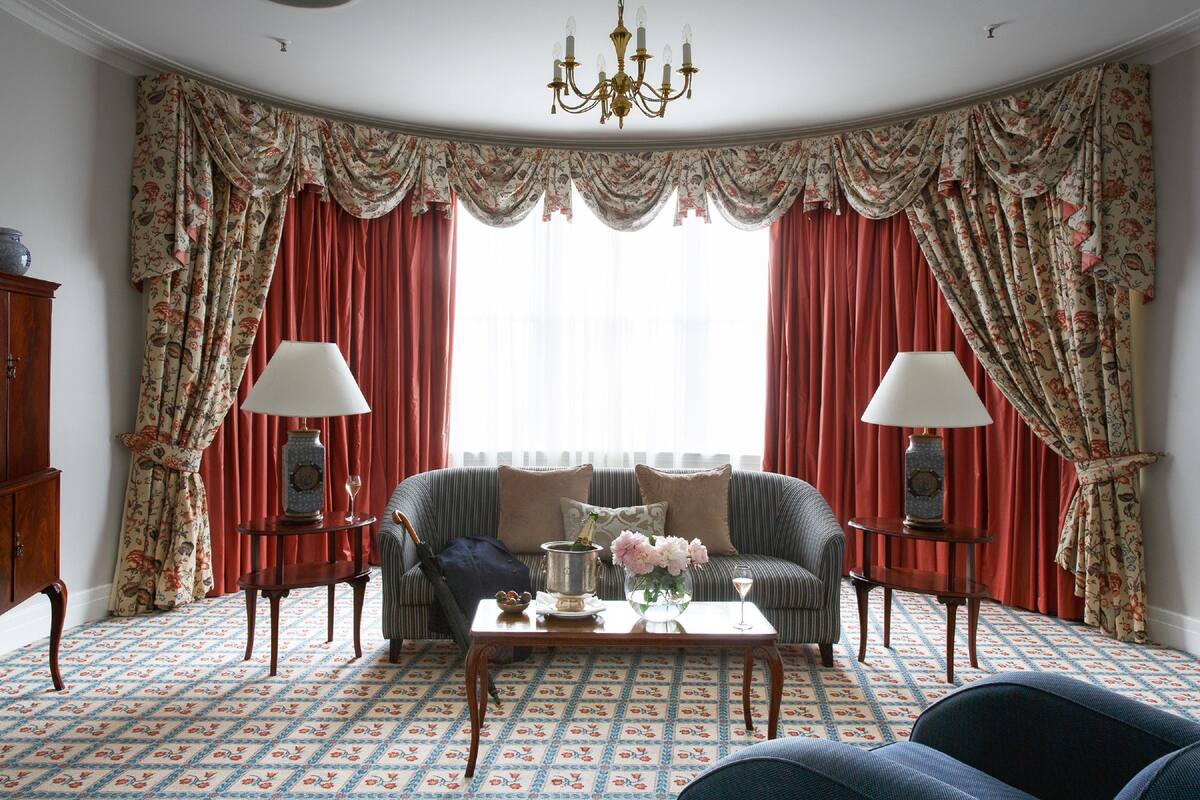 A room with a patterned carpet, a sofa, two side tables with lamps, a chandelier, and curtains with a floral pattern.
