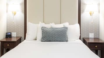 Una cama bien hecha con sábanas blancas y almohadas estampadas, con mesitas de noche a cada lado con lámparas y un reloj digital.