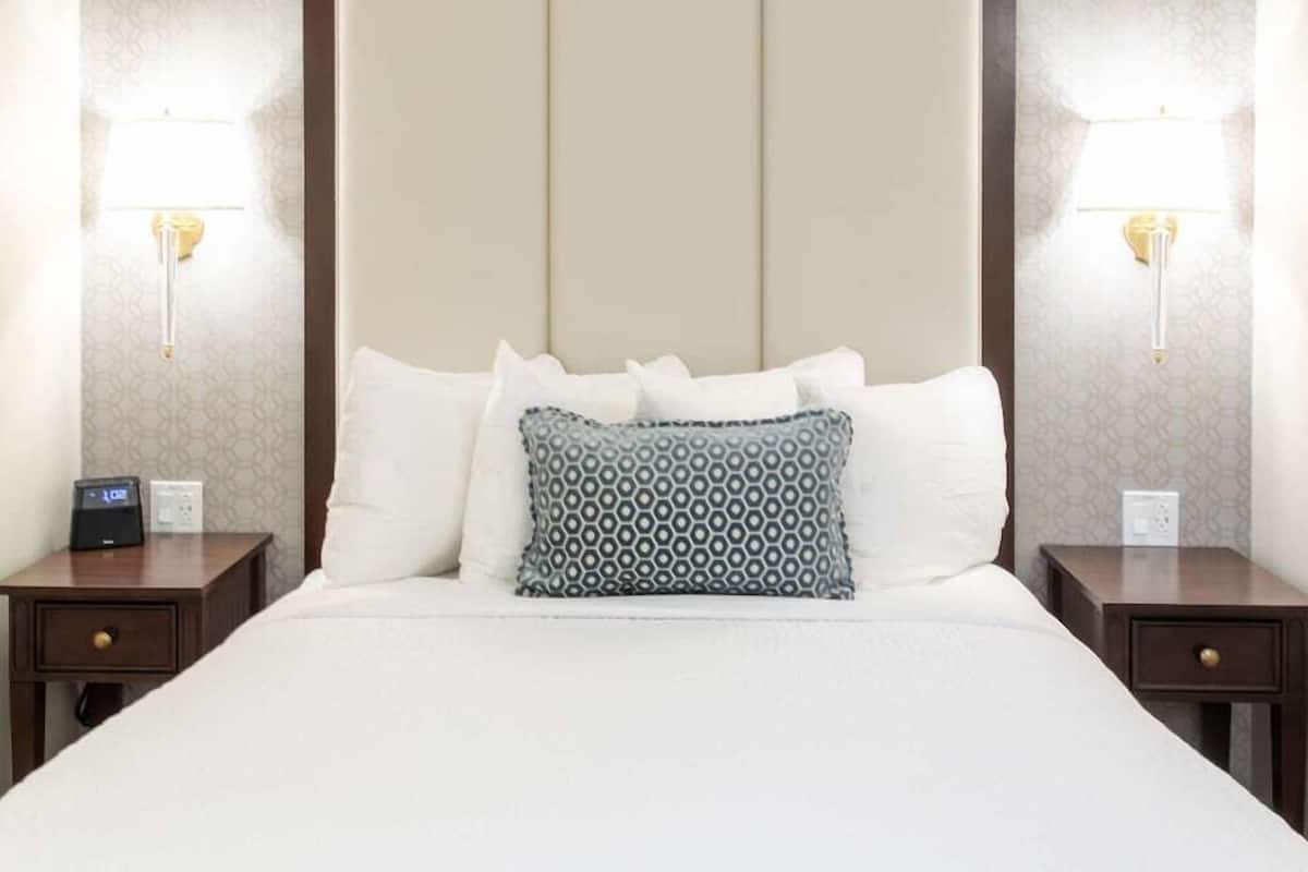 A hotel room with a large bed, two bedside tables, a nightstand, a digital clock, and two wall-mounted lamps.