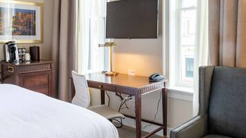 A hotel room with a bed, desk, chair, TV, and a view of the city.