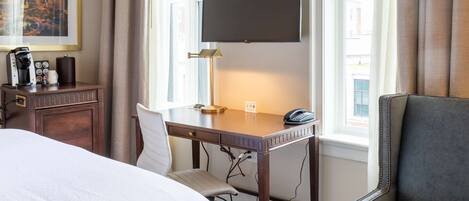A hotel room with a bed, desk, chair, TV, and a view of the city.
