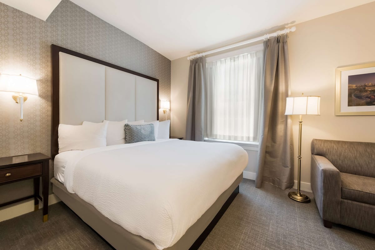 A hotel room with a large bed, a nightstand, a lamp, a chair, and a window with curtains.