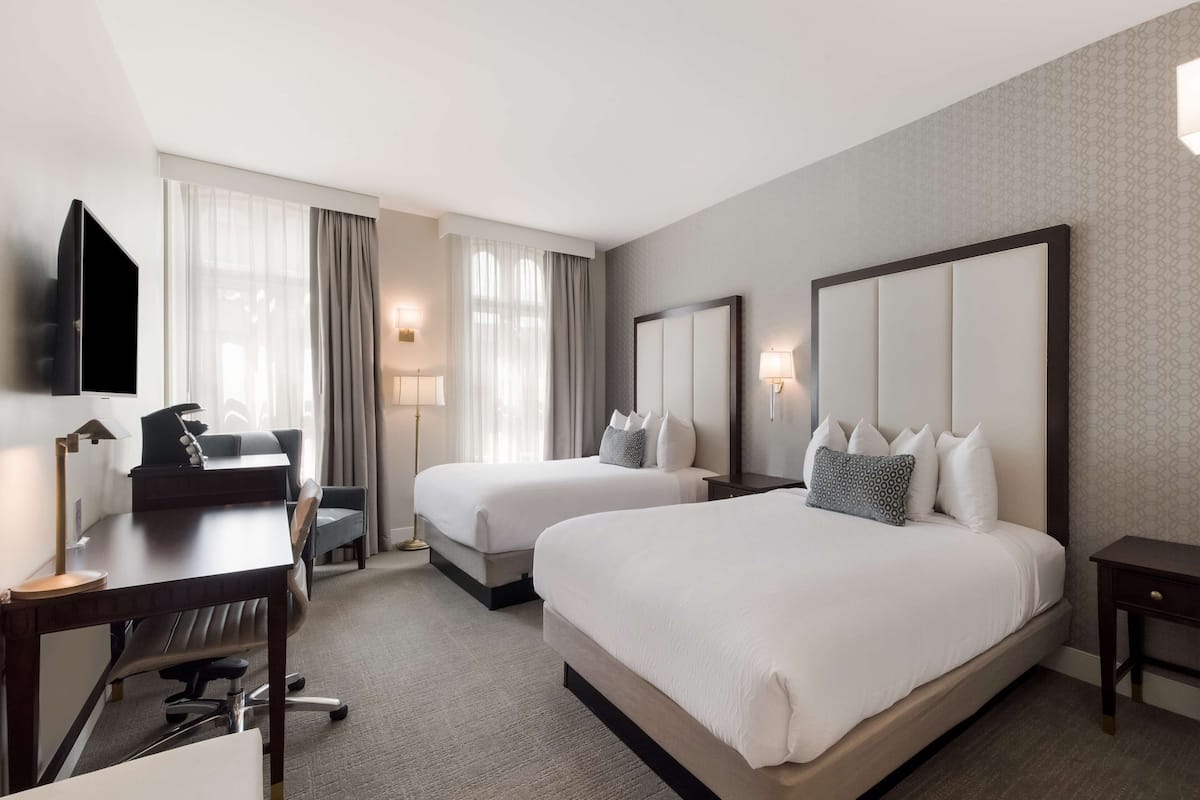 A hotel room with two beds, a desk with a chair, a TV, and a lamp.