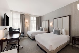 A hotel room with two beds, a desk with a chair, a TV, and a lamp.