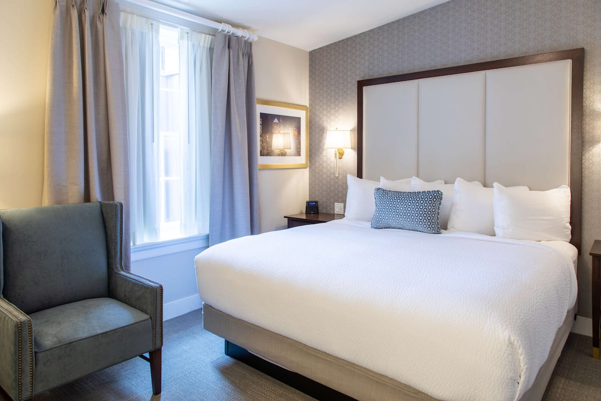 A hotel room with a large bed, a chair, a nightstand, a lamp, and a window with curtains.