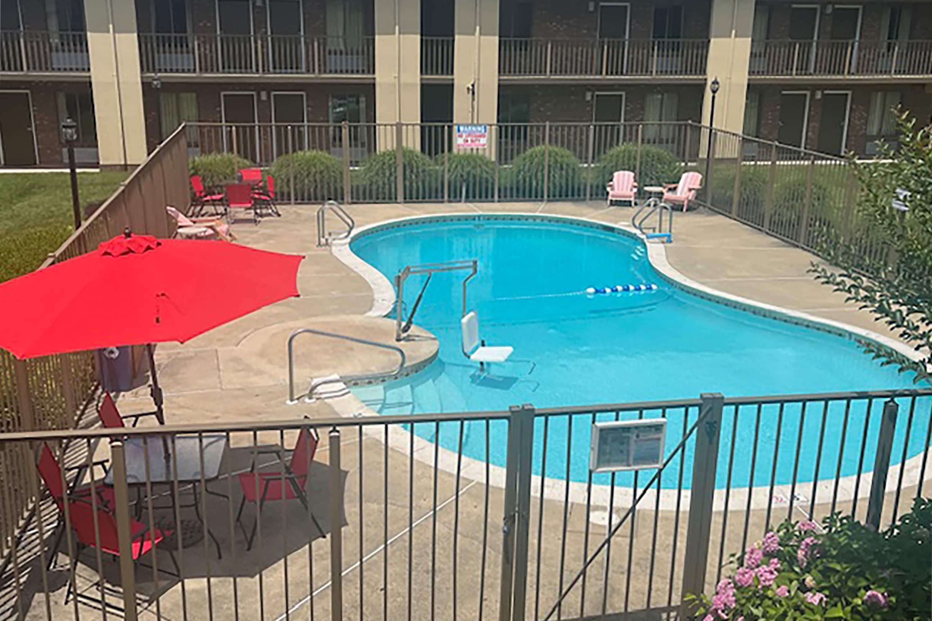 Une piscine avec un parasol rouge, des chaises longues et une zone délimitée par une clôture.