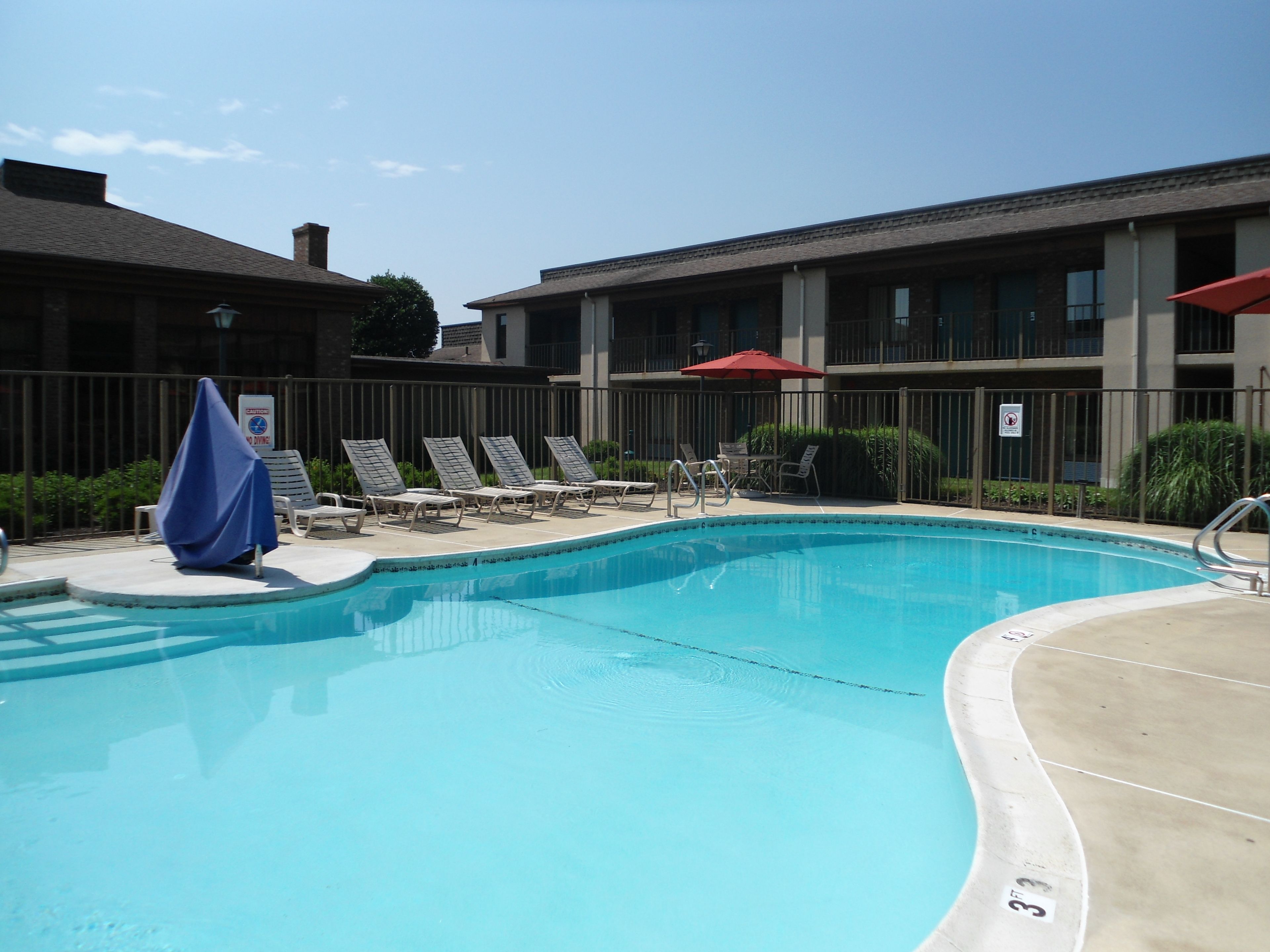A swimming pool with lounge chairs and umbrellas, surrounded by a fence and buildings.