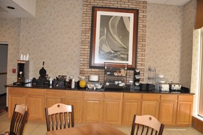 A hotel kitchen with a brick wall, a framed sailboat artwork, a coffee station, and wooden cabinets.